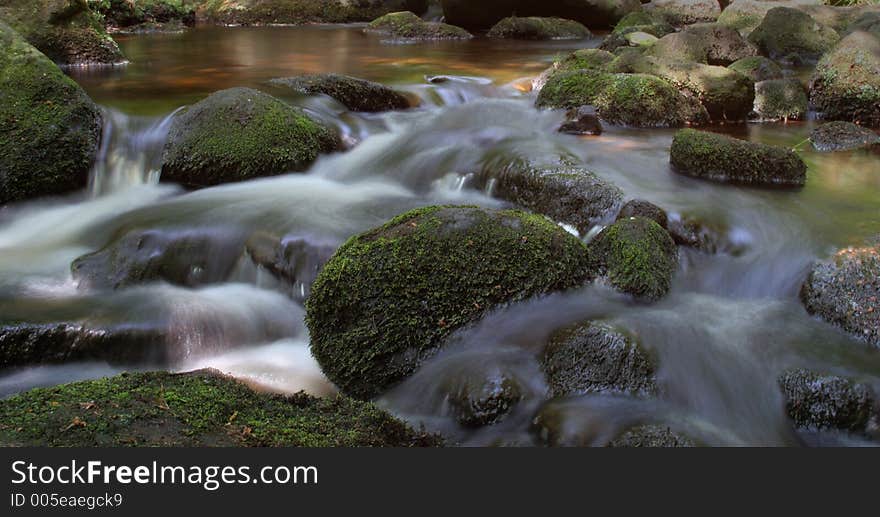 A wide shot of a stream with the motion of the water blurred around moss covered rocks and stones. A wide shot of a stream with the motion of the water blurred around moss covered rocks and stones