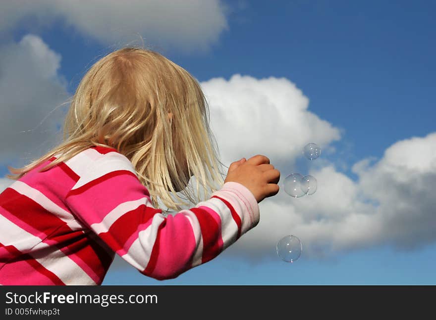 A little blond girl playing at blowing bubbles in sunny summer weather, set agains a blue sky with bubbles blowing away. A little blond girl playing at blowing bubbles in sunny summer weather, set agains a blue sky with bubbles blowing away