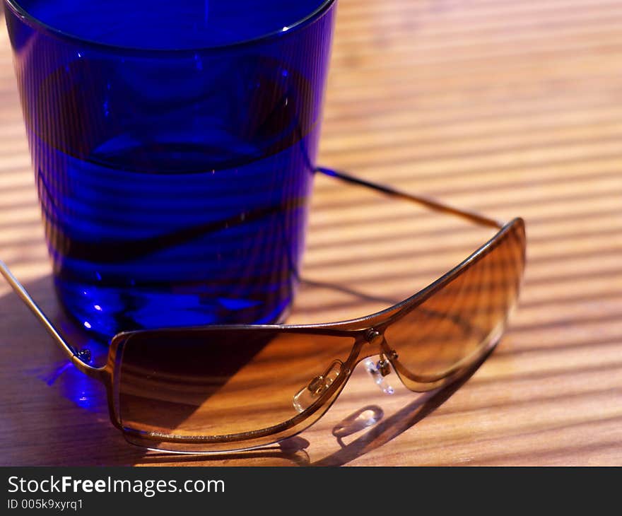 Womens' sunglasses resting next to a dark blue cocktail tumbler in strong light in shallow focus. Womens' sunglasses resting next to a dark blue cocktail tumbler in strong light in shallow focus.