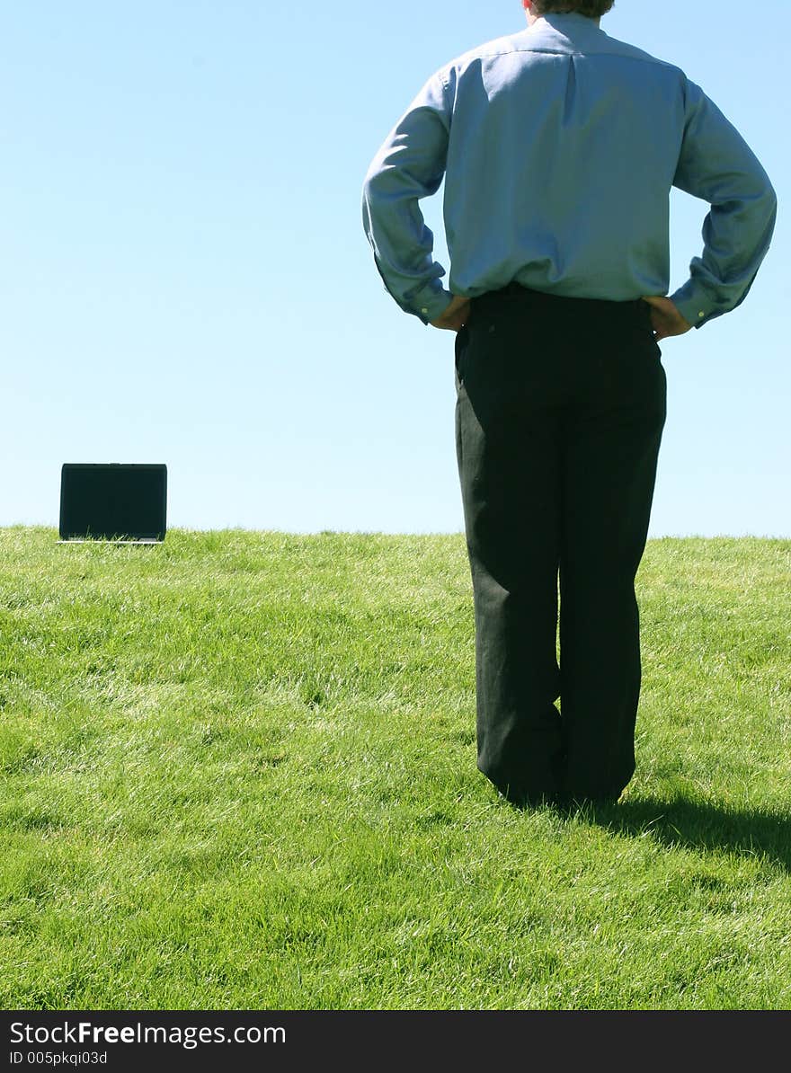 Business man in blue shirt standing holding his hands on his waist in the grass looking towards laptop that's in the grass. Business man in blue shirt standing holding his hands on his waist in the grass looking towards laptop that's in the grass