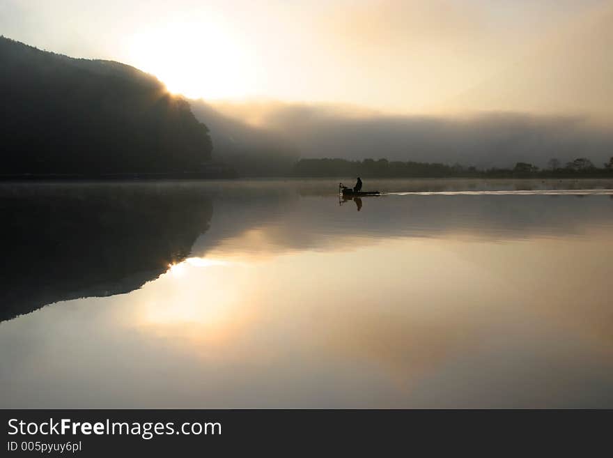 A lone fisherman on a tranquil and misty lake with the sun rising in the background. A lone fisherman on a tranquil and misty lake with the sun rising in the background.