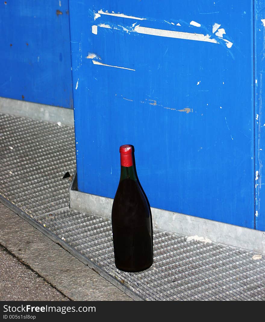 A full bottle with no label next to a blue rubbish bin. A full bottle with no label next to a blue rubbish bin