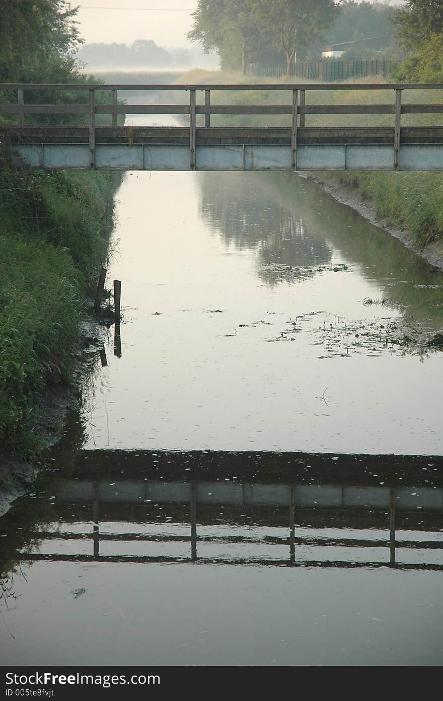 A bridge and its reflection crossing a still creek in the mist. A bridge and its reflection crossing a still creek in the mist.