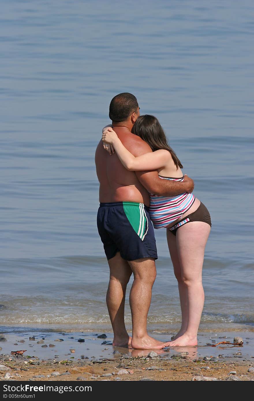 Mixed race couple at the beach of a pregnant female and her partner with their arms around each other, standing barefoot at the shoreline. Mixed race couple at the beach of a pregnant female and her partner with their arms around each other, standing barefoot at the shoreline.