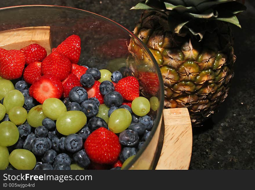 A bowl of fresh berries, strawberries, blueberries, grapes and a pineapple standing beside it. A bowl of fresh berries, strawberries, blueberries, grapes and a pineapple standing beside it.