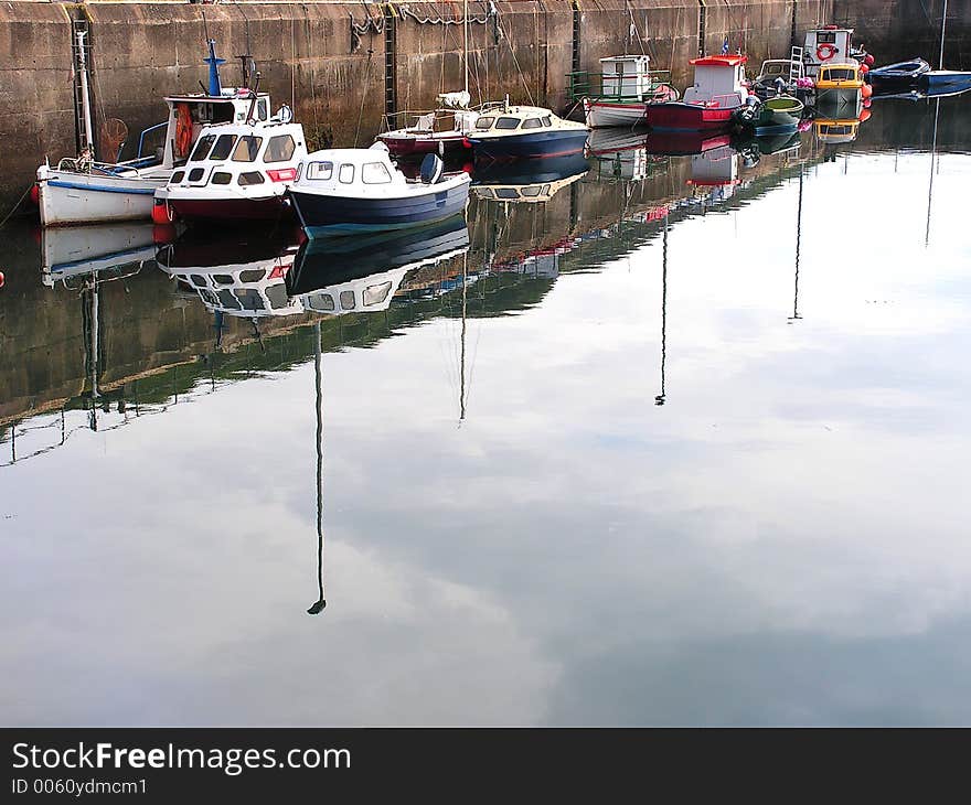 One side of the harbour with the summer season boats and yawls reflected in the calm still water with the sky. One side of the harbour with the summer season boats and yawls reflected in the calm still water with the sky.