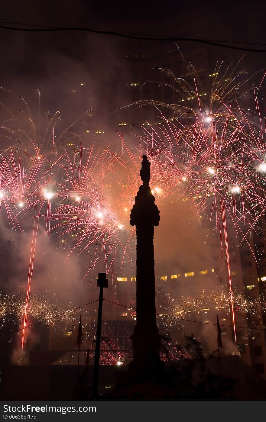 A fireworks display set off in a downtown setting between a skyscraper and a memorial. A fireworks display set off in a downtown setting between a skyscraper and a memorial