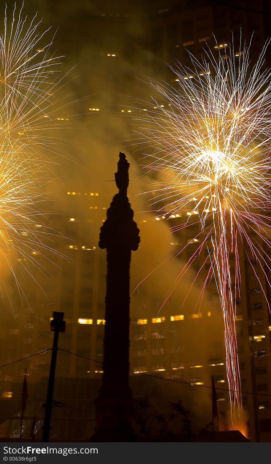 A fireworks display set off in a downtown setting with a skyscraper in the background and a memorial in the foreground. A fireworks display set off in a downtown setting with a skyscraper in the background and a memorial in the foreground