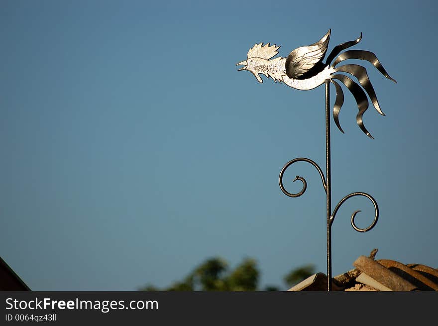 A metal chicken pictured on top of a roof, calling away, shining in the sun. A metal chicken pictured on top of a roof, calling away, shining in the sun.