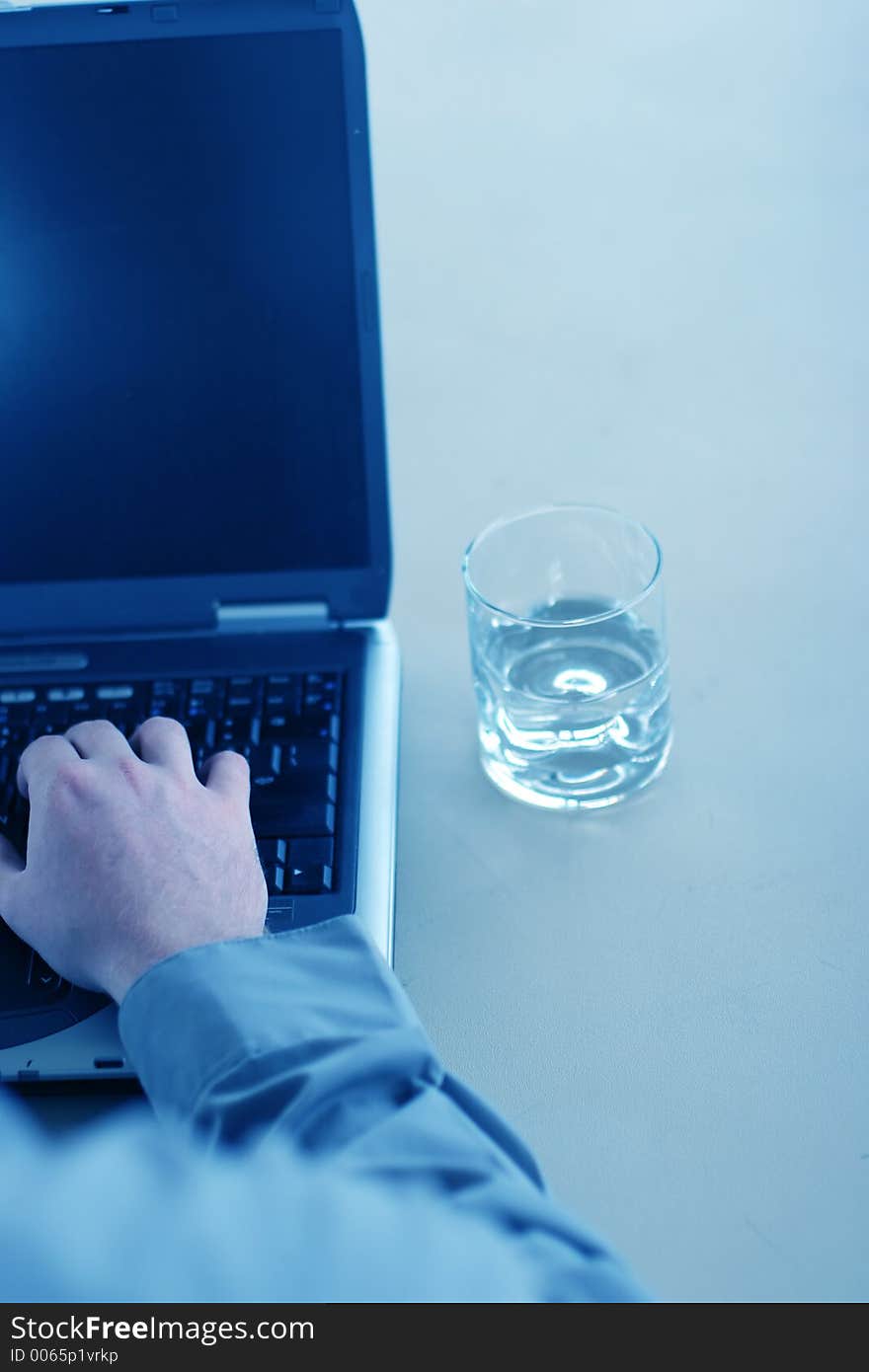 Business man in gray shirt is typing on his laptop with a glass of water next to him on the table. Business man in gray shirt is typing on his laptop with a glass of water next to him on the table