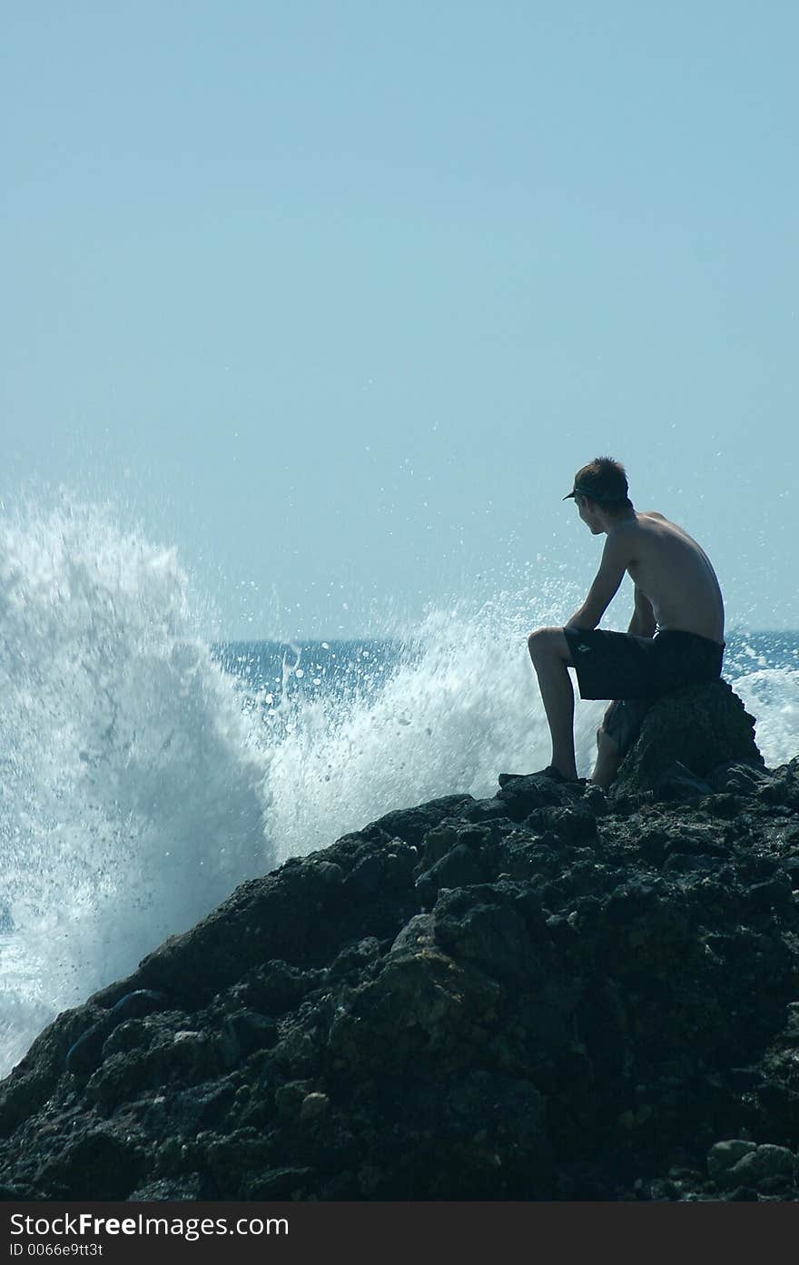 A boy is sitting on rocks and looking at the splashes of ocean waves. A boy is sitting on rocks and looking at the splashes of ocean waves