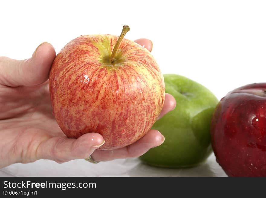 Hand holding one wet red apple with two apples, one red and one green, in background. Focus is one apple held in hand in the foreground. Hand holding one wet red apple with two apples, one red and one green, in background. Focus is one apple held in hand in the foreground.