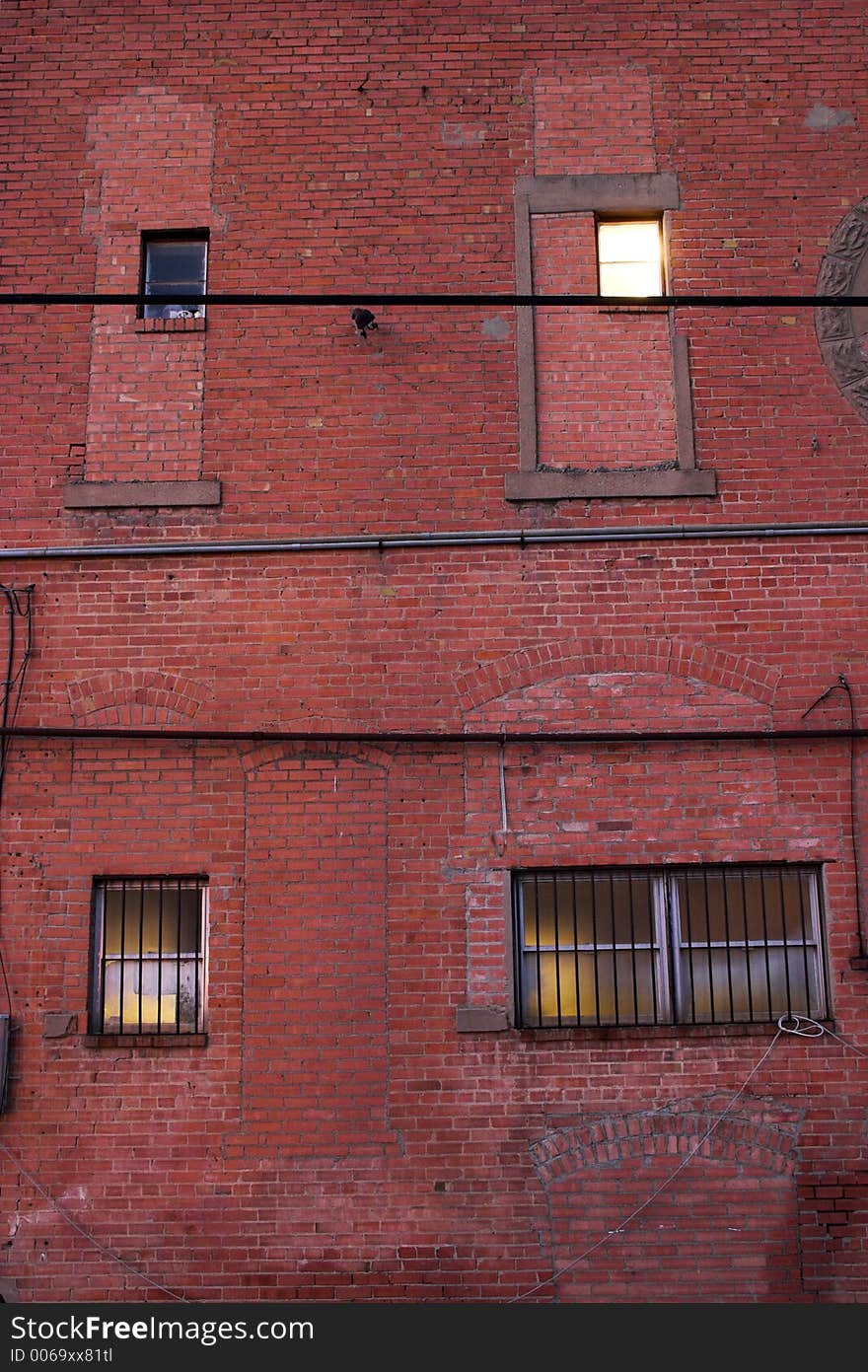 Four different windows on a grungy red brick wall in an alley. One upper window has a light brightly shining which contrasts with the dim glow from the other windows. Lower two windows have security bars. Four different windows on a grungy red brick wall in an alley. One upper window has a light brightly shining which contrasts with the dim glow from the other windows. Lower two windows have security bars.