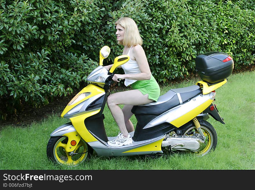 Beautiful and blond woman wearing white tank top and green shorts sitting on motor scooter in green grass. Beautiful and blond woman wearing white tank top and green shorts sitting on motor scooter in green grass.