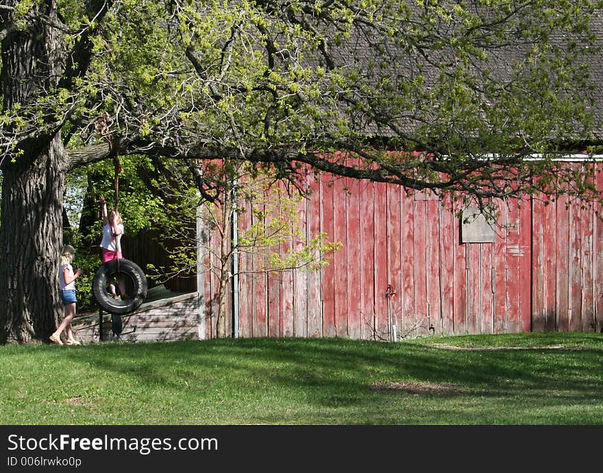 Two young girls playing on a tire swing on an oak tree with an old red barn in the background. Two young girls playing on a tire swing on an oak tree with an old red barn in the background.