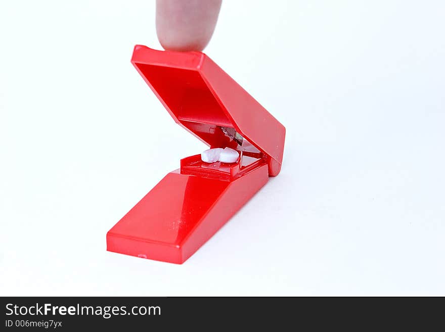 A red pill splitter which just split a white pill. The finger which pushed it is shown. White background.