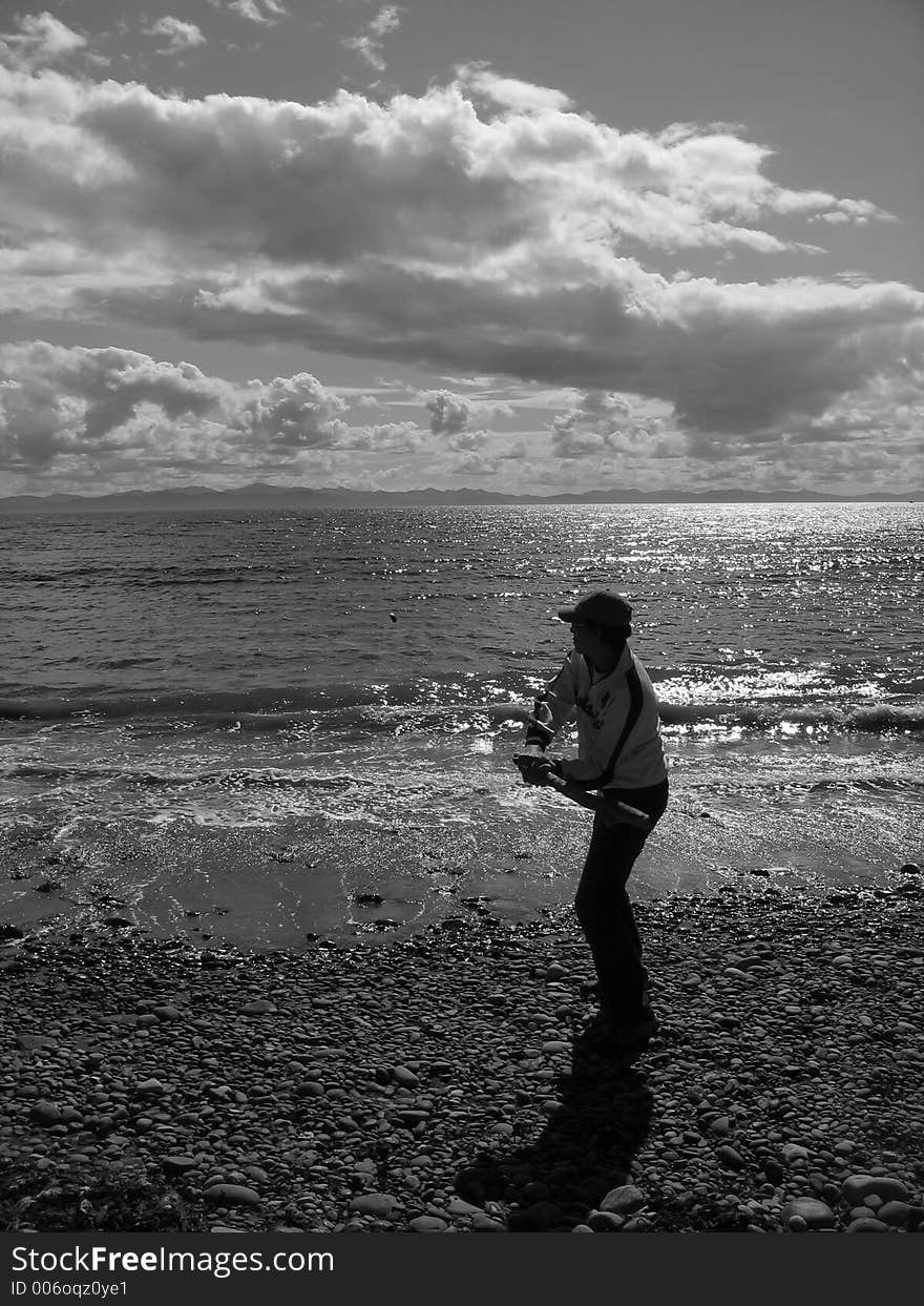 Man. standing at the beach hitting rocks with driftwood. Photo is in black and white with man in center ofpicture and ocean and interesting sky background. Man. standing at the beach hitting rocks with driftwood. Photo is in black and white with man in center ofpicture and ocean and interesting sky background