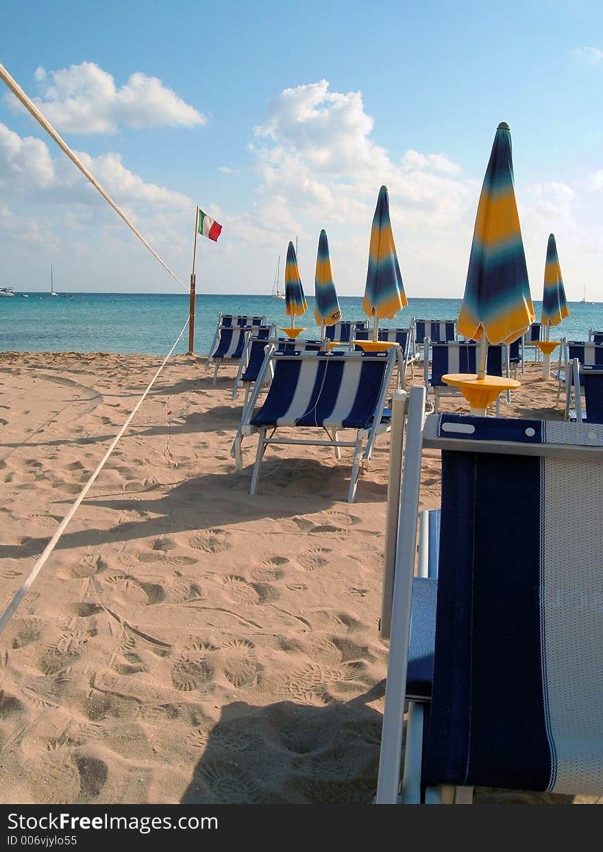 Row of closed beach umbrellas and deckchairs on a sicilian beach early in the morning. Row of closed beach umbrellas and deckchairs on a sicilian beach early in the morning