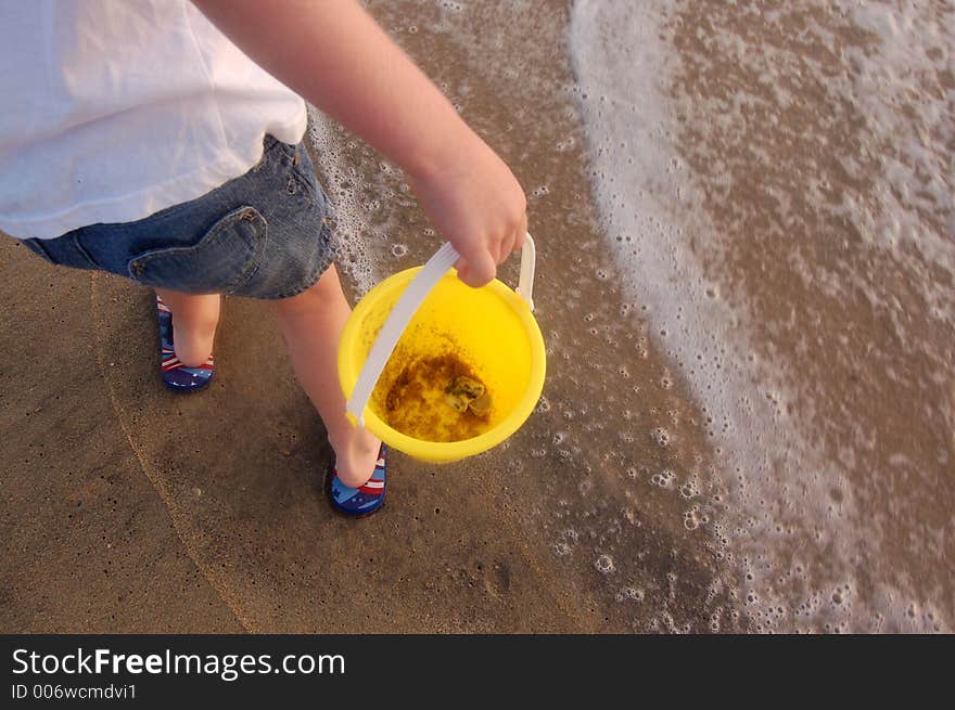 A child just showing her arm and hand carrying a yellow sand bucket on the beach,standing in the foamy waves. A child just showing her arm and hand carrying a yellow sand bucket on the beach,standing in the foamy waves.