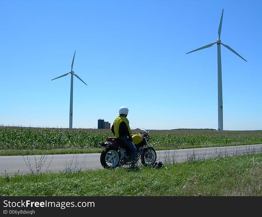 Motorcycle Rider rests while taking in sights of towering windmills. Wearing helmet? Of course. Motorcycle Rider rests while taking in sights of towering windmills. Wearing helmet? Of course.