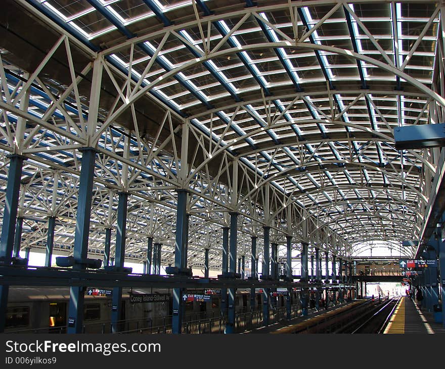 This is the architecture of a large outdoor subway terminal. Very graphic, the cieling and sides are shown in daylight. The ceiling is curved and open with a grid design. The stanchions are painted blue