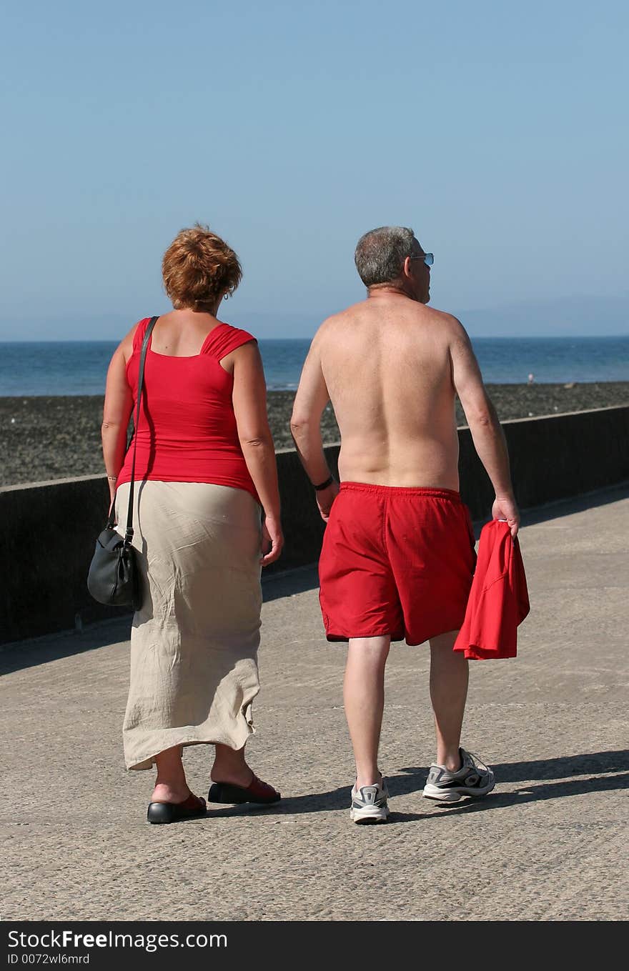 Rear view of a middle aged couple both wearing red, walking together along a beach promenade with the sea and a blue sky to the rear. Rear view of a middle aged couple both wearing red, walking together along a beach promenade with the sea and a blue sky to the rear.