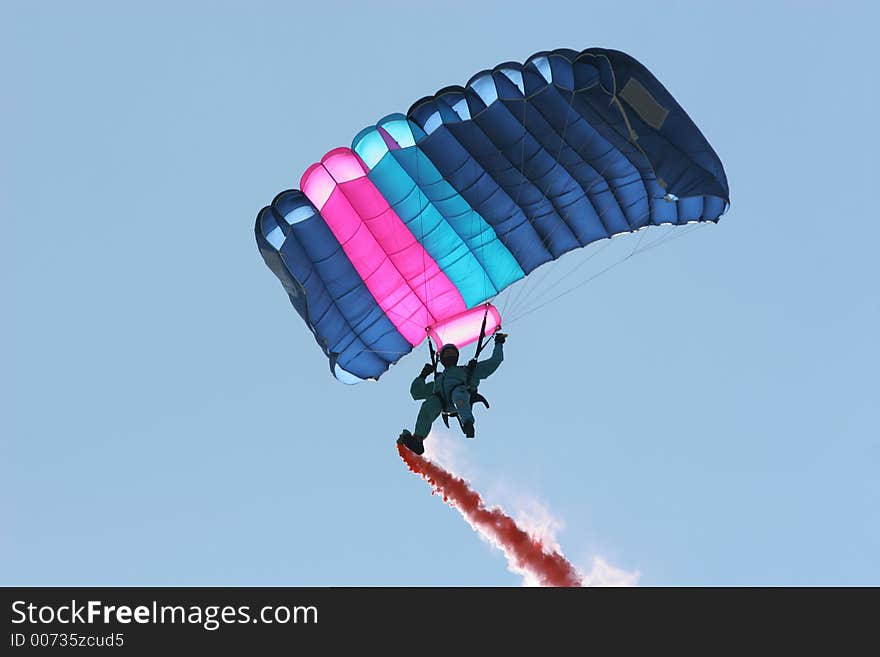 Parachutist in a blue boiler suit, with a pink and blue parachute, flying through the air with a red smoke trail coming from his right foot. Parachutist in a blue boiler suit, with a pink and blue parachute, flying through the air with a red smoke trail coming from his right foot..