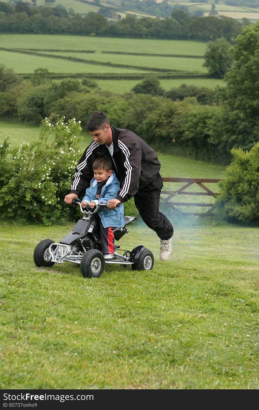 Little boy on racing a mini moto quad bike with his father riding on the back. Set in countryside. Little boy on racing a mini moto quad bike with his father riding on the back. Set in countryside.