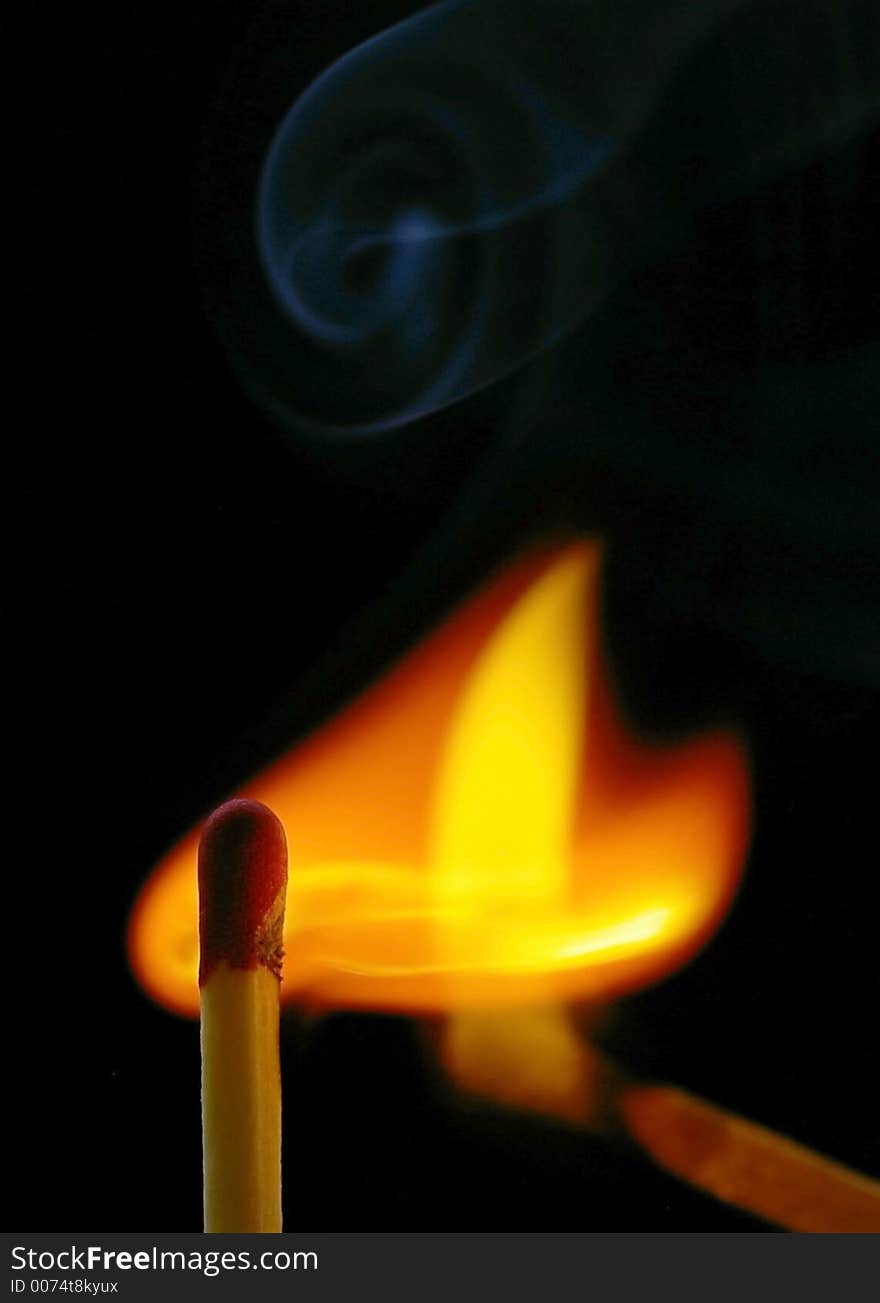 Closeup of a red-tipped wooden match stick at ignition from another match.  Red, orange, yellow and white flame.  Visible wift of Smoke.  Black background. Closeup of a red-tipped wooden match stick at ignition from another match.  Red, orange, yellow and white flame.  Visible wift of Smoke.  Black background.