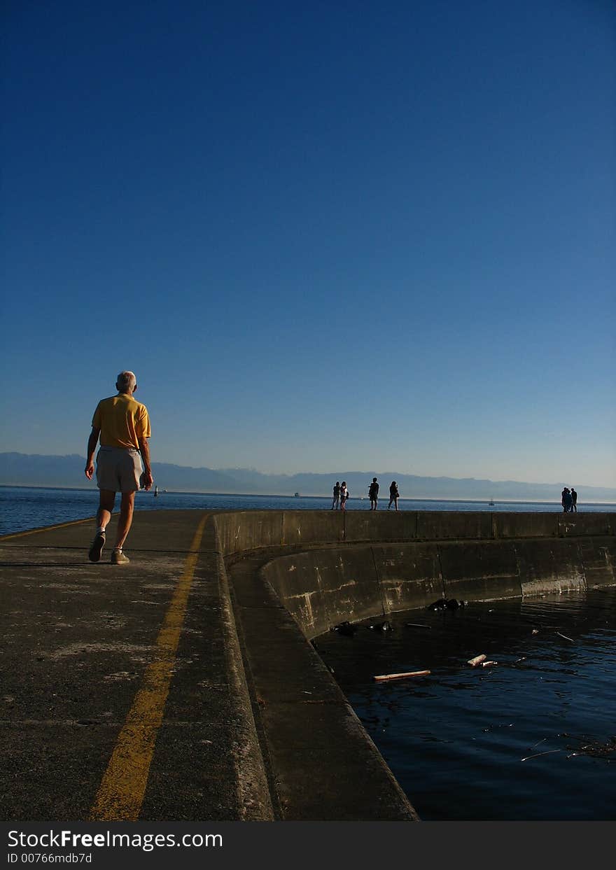 Man walking along seaside pier with peir leading off into distance.  Other walkers can also be seen. Sky overhead is rich blue and photo is shot vertically. Man walking along seaside pier with peir leading off into distance.  Other walkers can also be seen. Sky overhead is rich blue and photo is shot vertically