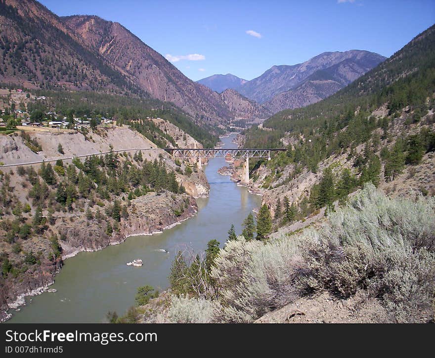 View of mountain river with train trestle and small town, mountains in background.