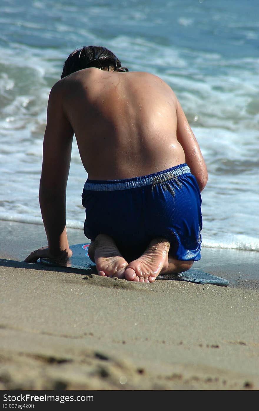 A boy is sitting on the skim board in the beach and 
looking for something in the sand. A boy is sitting on the skim board in the beach and 
looking for something in the sand