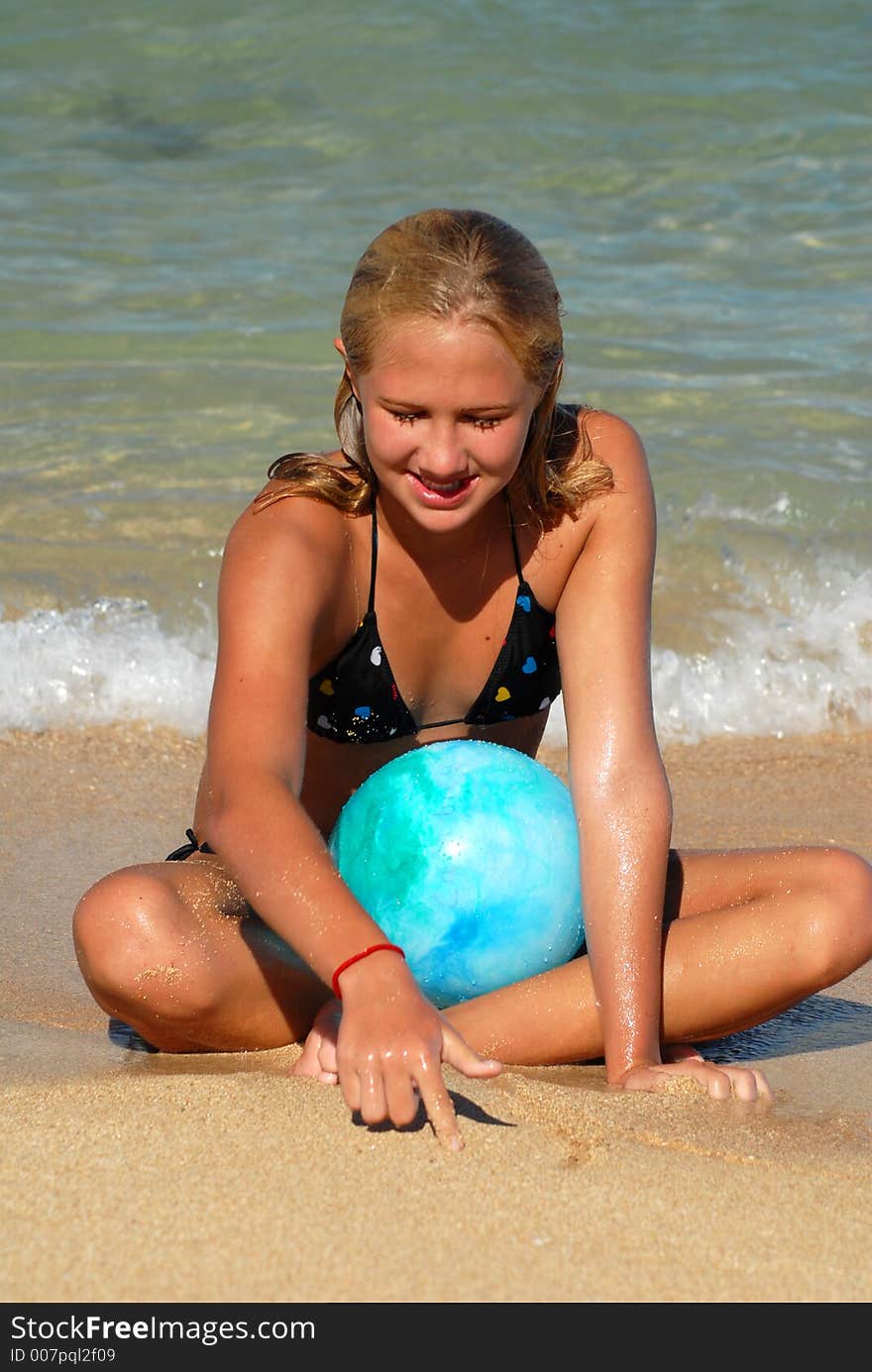 A teenager girl with a blue ball is sitting on a sandy beach near the water. A teenager girl with a blue ball is sitting on a sandy beach near the water