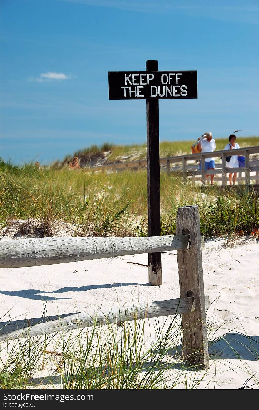 Beach scene with fence and sign that says Keep off the Dunes. There is sand, grass and a blue sky. There are people visible in the blurred background. Beach scene with fence and sign that says Keep off the Dunes. There is sand, grass and a blue sky. There are people visible in the blurred background.