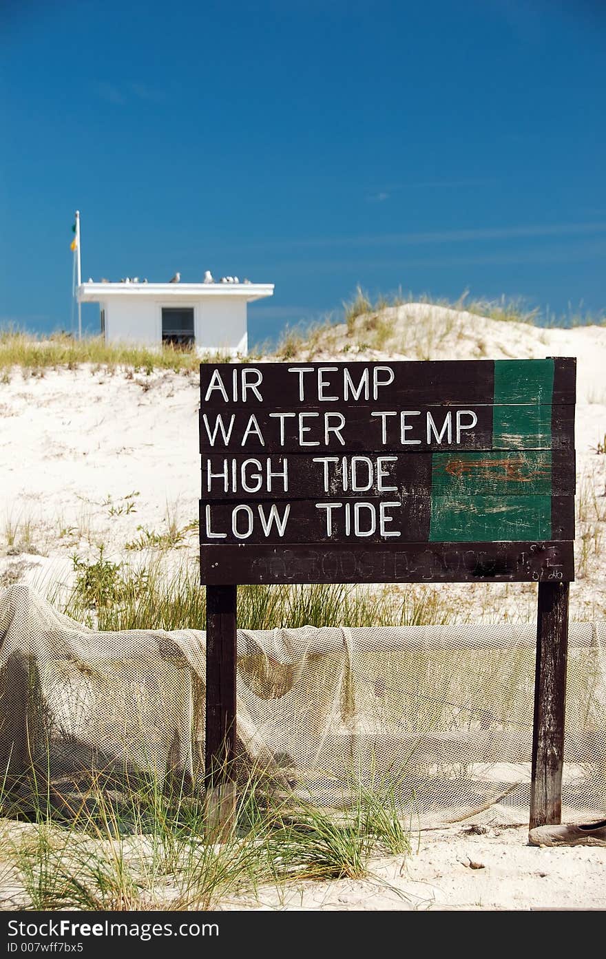 A beach scene with a fence, net and water conditions sign.  A lifeguard shack, flags and seagulls are in blurred background.  The sky is blue almost cloudless. A beach scene with a fence, net and water conditions sign.  A lifeguard shack, flags and seagulls are in blurred background.  The sky is blue almost cloudless.