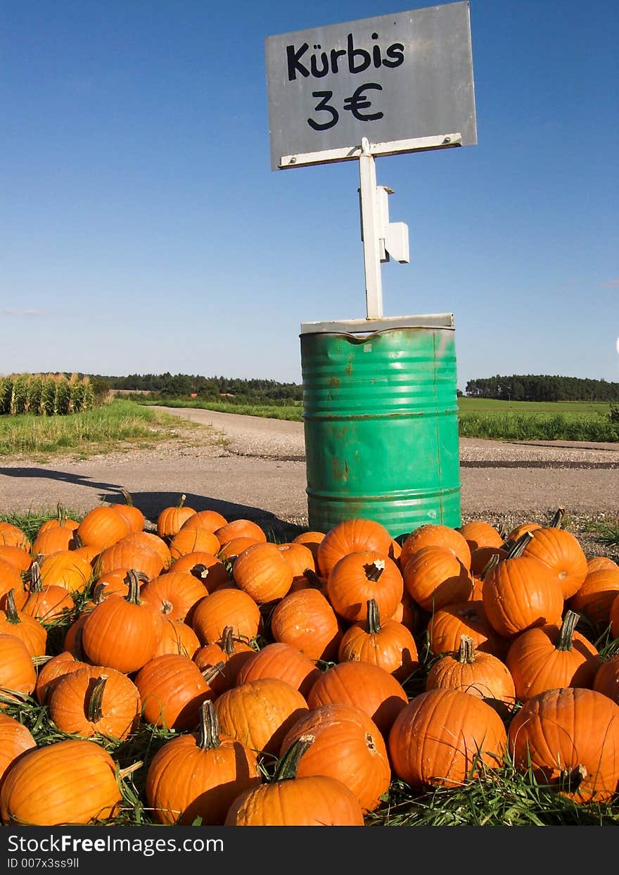 The image shows a pile of orange pumpkins (in German it is 'KÃ¼rbis') before a green barrel. On top of the barrel is a silver sign, showing that a pumpkin costs 3 Euro. Behind the barrel is a small road. The sky in the picture is blue. The cucurbits are mainly sold as halloween decoration. The image shows a pile of orange pumpkins (in German it is 'KÃ¼rbis') before a green barrel. On top of the barrel is a silver sign, showing that a pumpkin costs 3 Euro. Behind the barrel is a small road. The sky in the picture is blue. The cucurbits are mainly sold as halloween decoration.