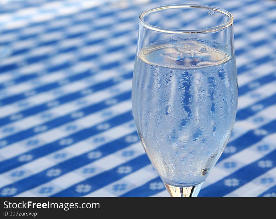 A glass of iced water, standing on a blue and white tablecloth, ice cubes in glass and condensed water on glass due to heat. A glass of iced water, standing on a blue and white tablecloth, ice cubes in glass and condensed water on glass due to heat