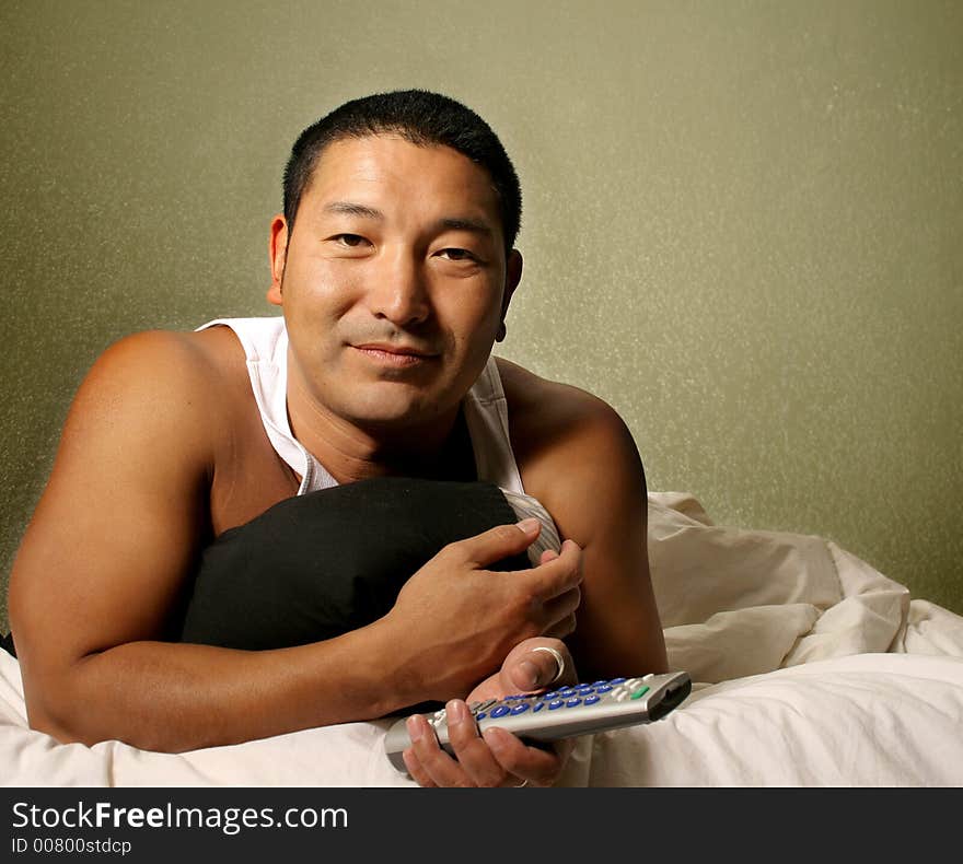 Asian man laying in bed holding a remote control. Asian man laying in bed holding a remote control