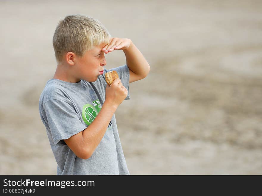 Young boy is watching the scenery at the beach and is eating a hamburger. Young boy is watching the scenery at the beach and is eating a hamburger