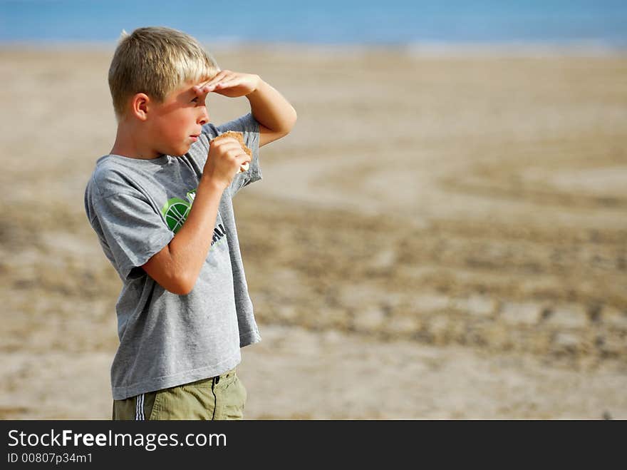 Young boy is watching the scenery at the beach and is eating a hamburger. Young boy is watching the scenery at the beach and is eating a hamburger