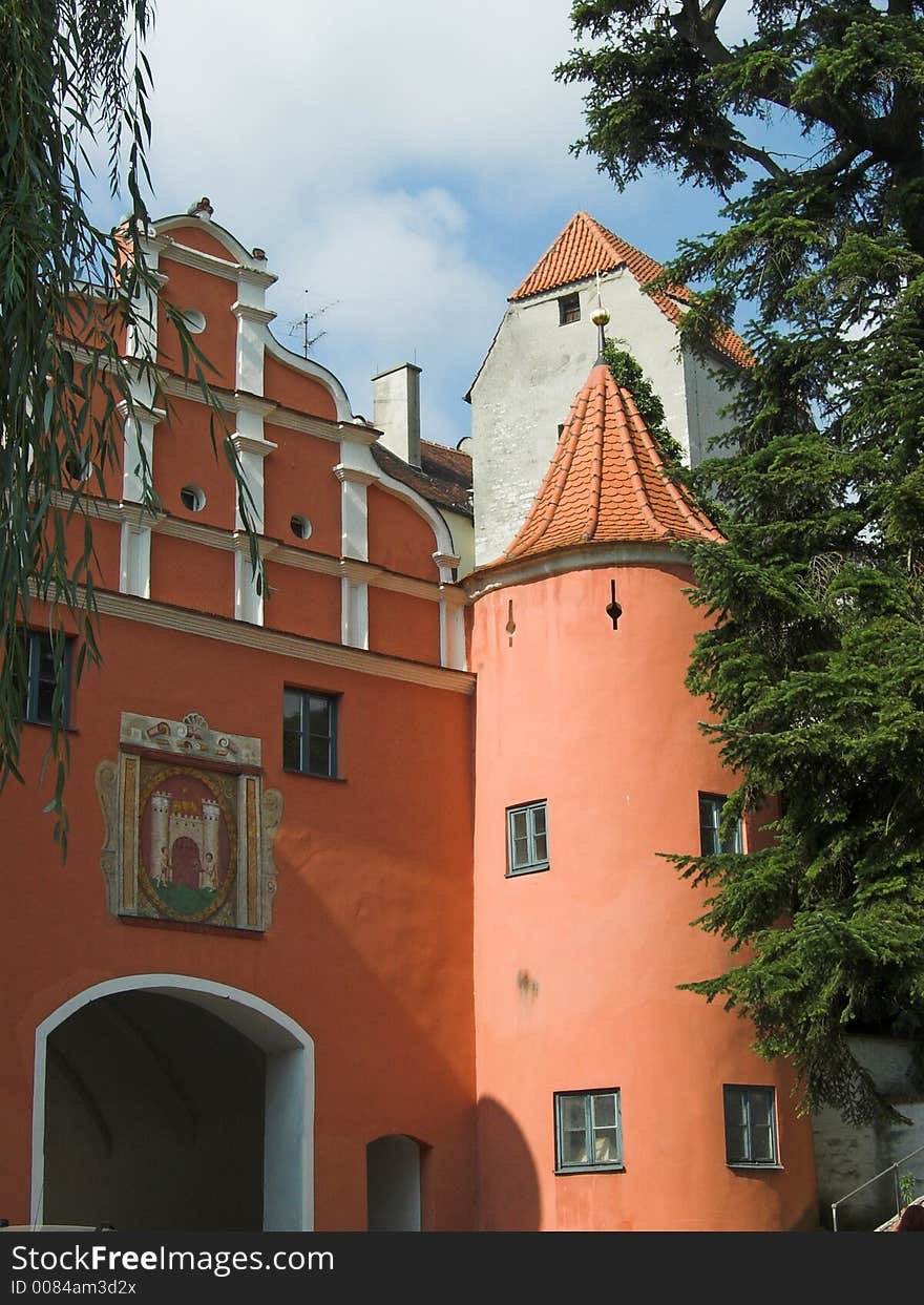 The picture shows a old, historic city gate in the town of Neuburg (Germany, Bavaria). It is a read building with an icon above the door. On the right side of the gate is a tower. The image is framed by two trees.