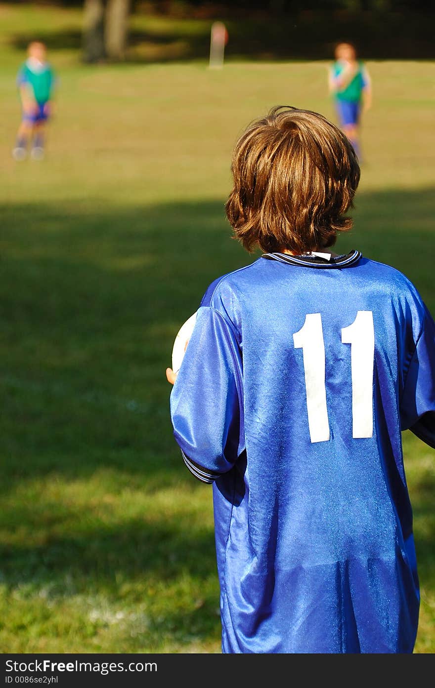 Young boy playing soccer on s sunny afternoon. His back is to the camera. There are two other players visible in the blurred background. Young boy playing soccer on s sunny afternoon. His back is to the camera. There are two other players visible in the blurred background.