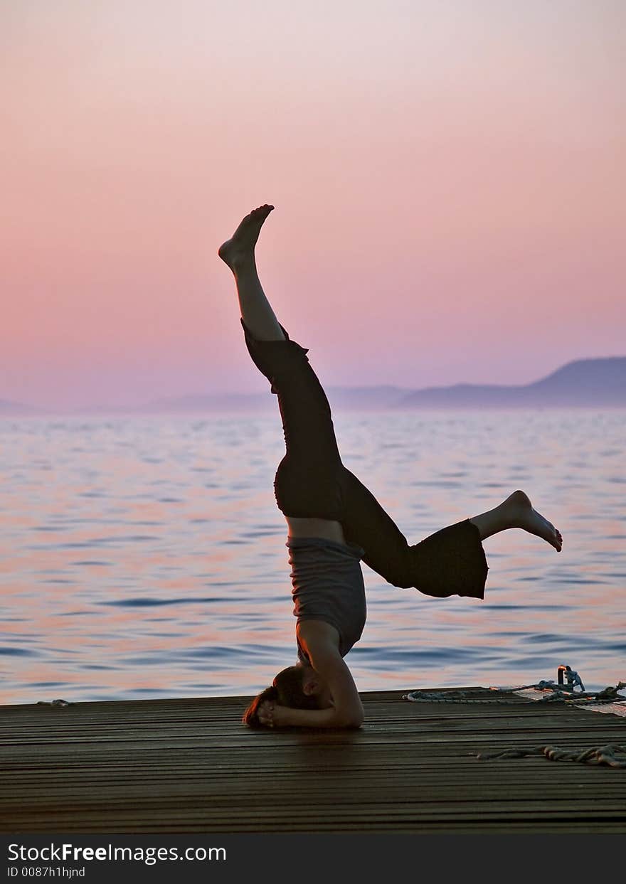 Young woman exercising yoga, balancing on the head, sea and sunset at the background. Young woman exercising yoga, balancing on the head, sea and sunset at the background