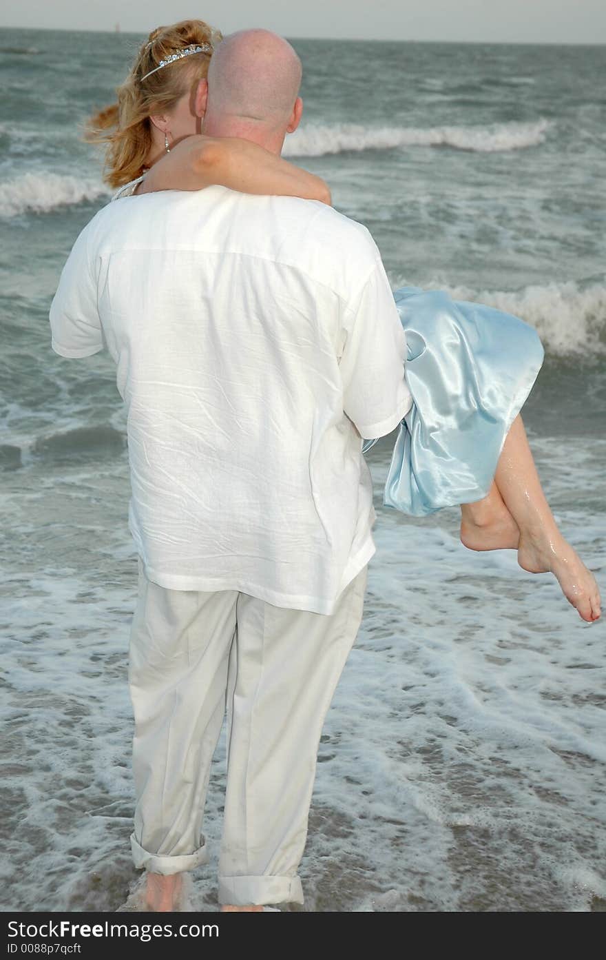 Happy groom picks up his bride. Man holding his woman in his arms on the beach shore. Standing in the waves of the ocean. Couple facing the ocean. Happy groom picks up his bride. Man holding his woman in his arms on the beach shore. Standing in the waves of the ocean. Couple facing the ocean.