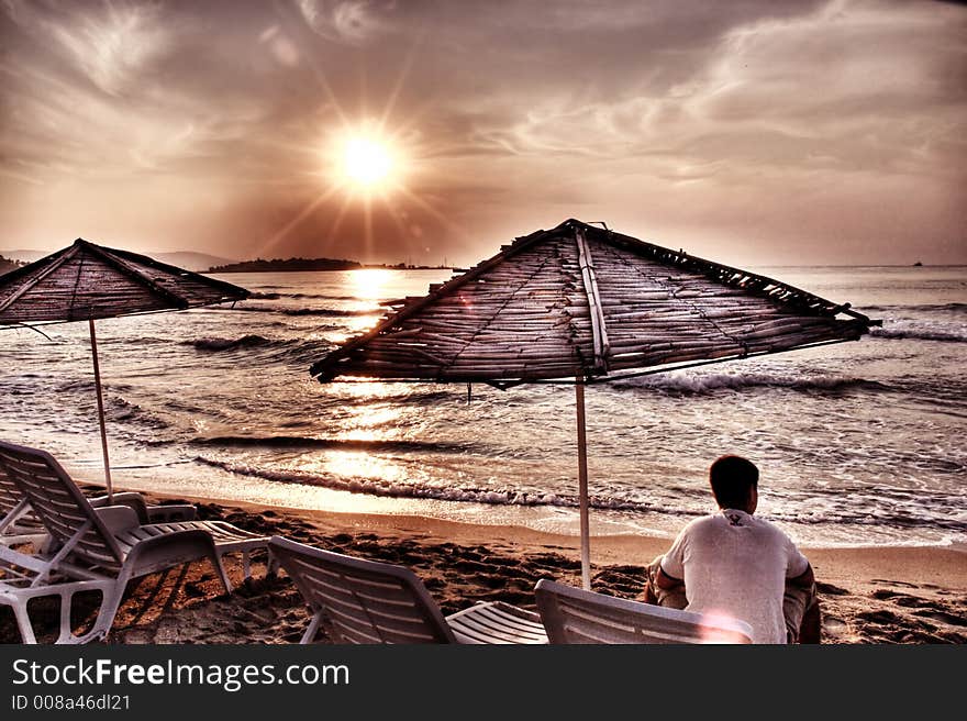 Man sitting in lounge under sunshade and looking at the sea at sunrise. Artistic photo. Noice visible at 100%. This is HDR photo with vivid and warm colors. Man sitting in lounge under sunshade and looking at the sea at sunrise. Artistic photo. Noice visible at 100%. This is HDR photo with vivid and warm colors.