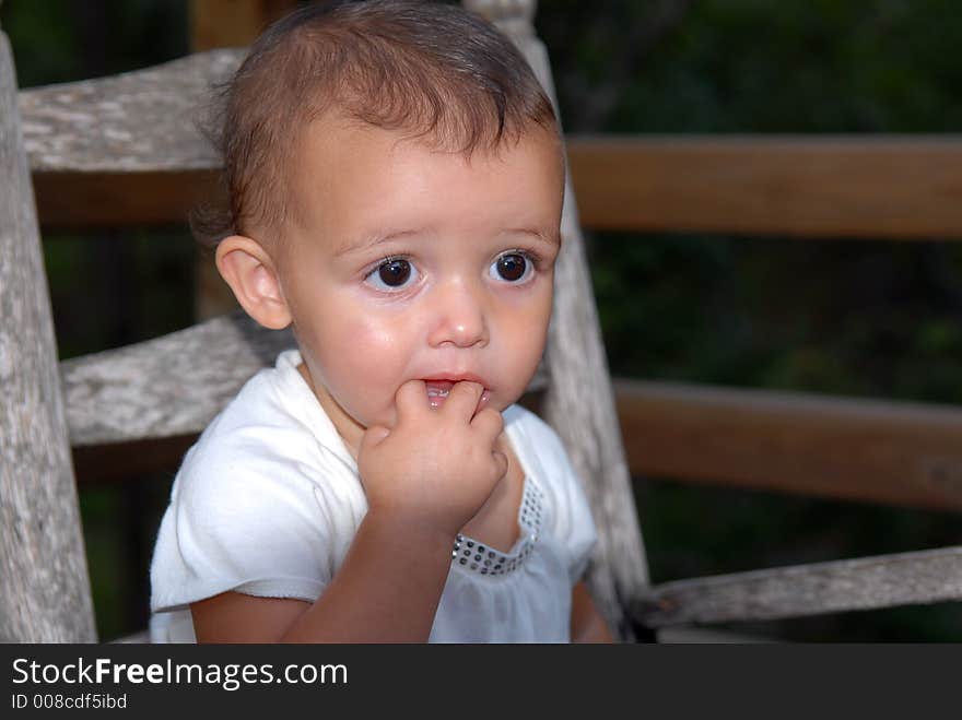 A beautiful little girl with big brown eyes and a sweet expression rocking in an old rocker with her fingers in her mouth. A beautiful little girl with big brown eyes and a sweet expression rocking in an old rocker with her fingers in her mouth