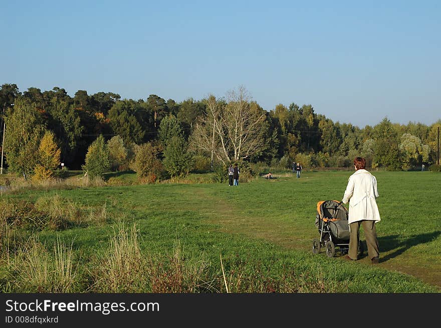 Autumn park. Trees still staying green with some golden colours. Close to camera: mother pushing her baby`s carriage.