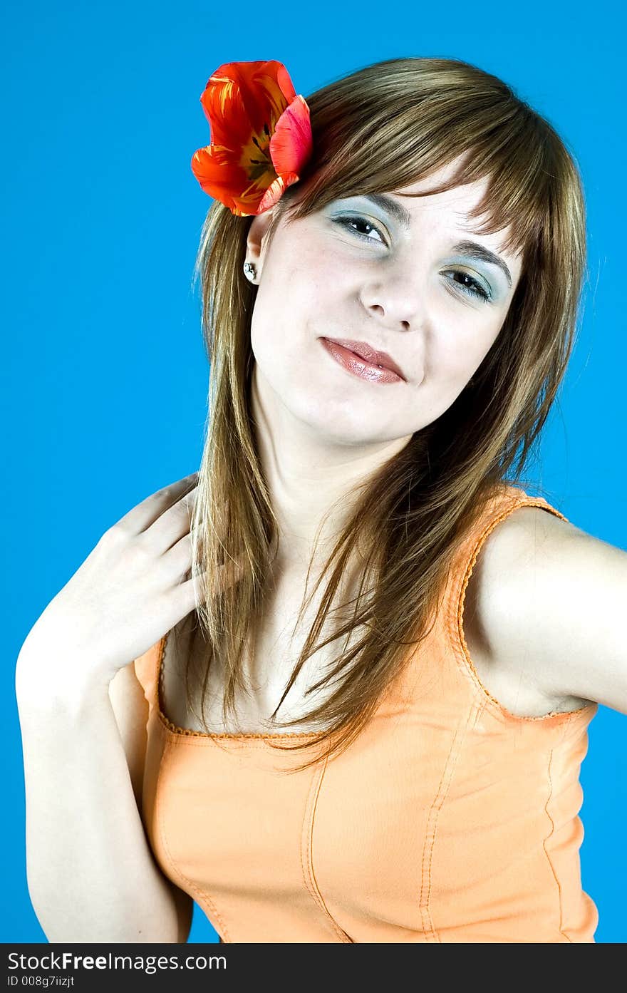 Portrait of a young girl smiling, with a red tulip over her ear, looking confident ih her youth and looks; nice make-up; posing in a studio; expression of youth; isolated on blue background. Portrait of a young girl smiling, with a red tulip over her ear, looking confident ih her youth and looks; nice make-up; posing in a studio; expression of youth; isolated on blue background