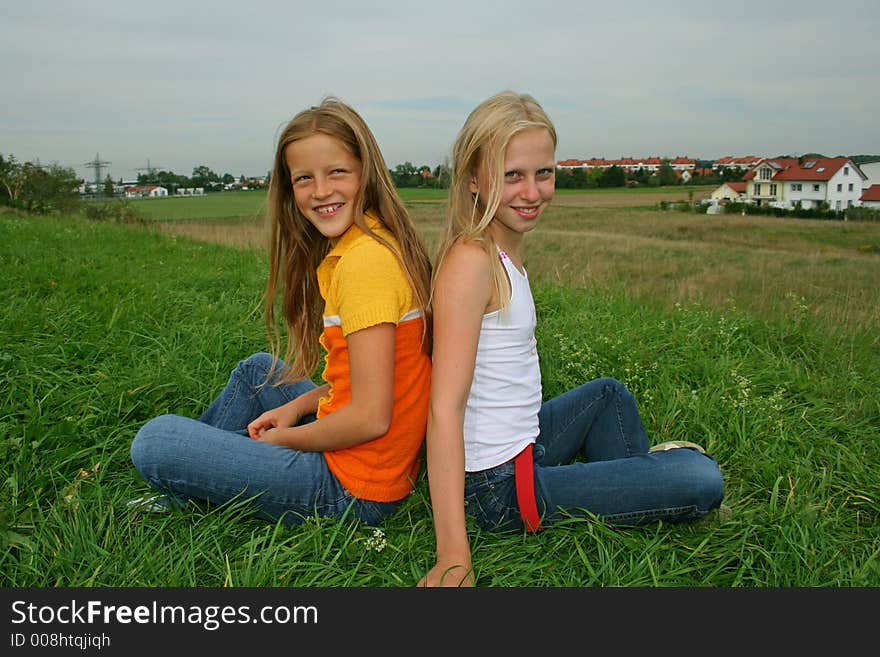 Two younf girl friends sitting in cross-legged pose in field. Two younf girl friends sitting in cross-legged pose in field