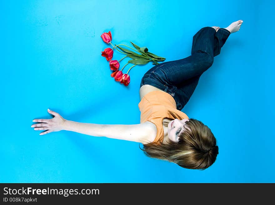 Portrait of a young girl laying on the floor and looking at a bouquet of red tulips and smiling, expression of youth and happiness; nice make-up, isolated on blue background. Portrait of a young girl laying on the floor and looking at a bouquet of red tulips and smiling, expression of youth and happiness; nice make-up, isolated on blue background